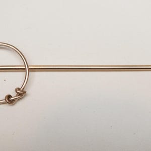 Op de afbeelding: Goudkleurige haarspeld met een cirkelvormig ontwerp en een lange, puntige pin. Het cirkelvormige gedeelte heeft een decoratief knoopontwerp. Een eenvoudig, elegant accessoire.