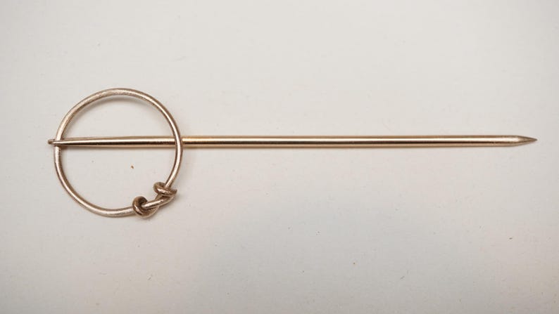 Op de afbeelding: Goudkleurige haarspeld met een cirkelvormig ontwerp en een lange, puntige pin. Het cirkelvormige gedeelte heeft een decoratief knoopontwerp. Een eenvoudig, elegant accessoire.