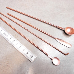 May include: Four copper cocktail stirring tools, including a spoon, a flat stirring tool, a twisted stirring tool, and a spoon with a rounded end. The tools are laid out on a grey surface with a ruler in the background.
