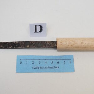 Puede incluir: Un cuchillo de manualidades con mango de madera clara y hoja de metal oscuro y moteado con una punta afilada y angular. Un peque&ntilde;o letrero blanco con la letra D se sit&uacute;a sobre la hoja, junto a una regla azul con escala en cent&iacute;metros.