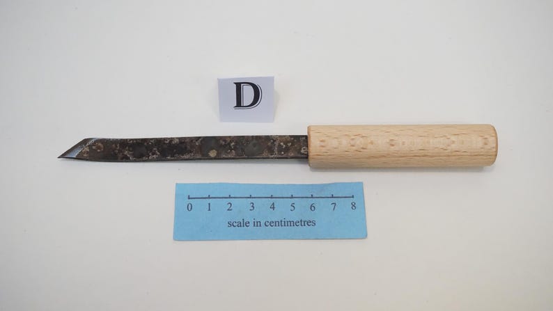 Puede incluir: Un cuchillo de manualidades con mango de madera clara y hoja de metal oscuro y moteado con una punta afilada y angular. Un peque&ntilde;o letrero blanco con la letra D se sit&uacute;a sobre la hoja, junto a una regla azul con escala en cent&iacute;metros.