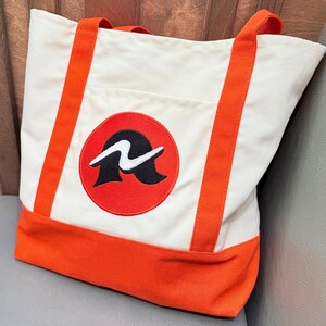 Peut inclure: Sac fourre-tout en toile écrue avec des détails orange. Il présente une base orange, des sangles assorties et un emblème circulaire avec un motif noir et blanc. Une poche est visible à l'avant.