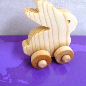 May include: A wooden toy bunny on wheels. The bunny is made of light-colored wood with visible grain, and has a cut-out design for the ears. The wheels are a contrasting light brown color. The toy is set against a purple background.