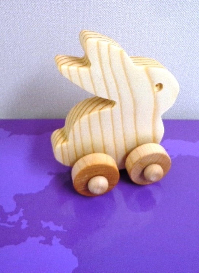 May include: A wooden toy bunny on wheels. The bunny is made of light-colored wood with visible grain, and has a cut-out design for the ears. The wheels are a contrasting light brown color. The toy is set against a purple background.