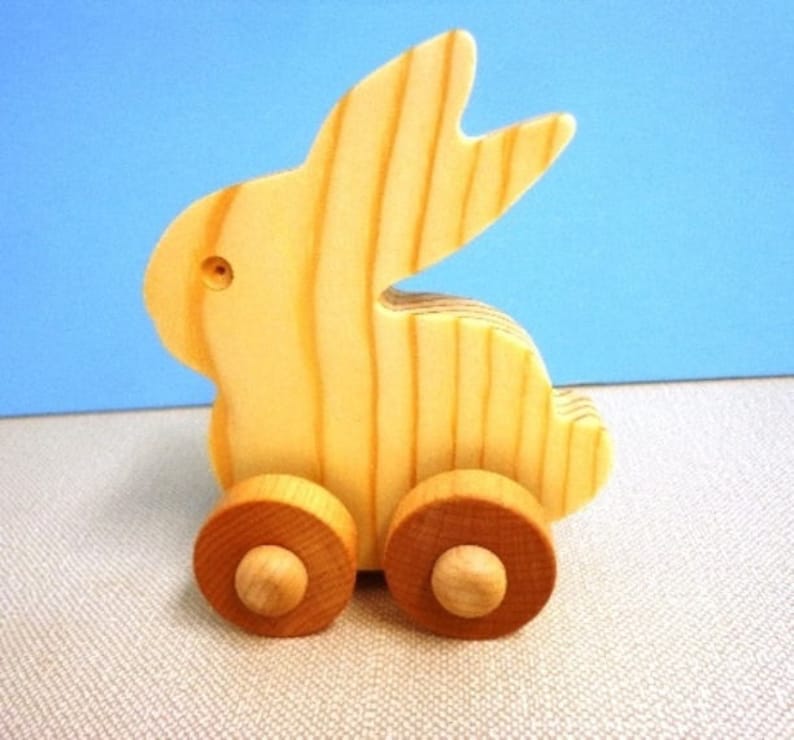 May include: A wooden toy bunny on wheels. The bunny is made of light-colored wood with visible grain, and has large ears. The wheels are brown and attached to the bunny's body. The toy is designed for children.