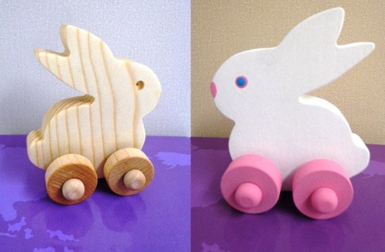 May include: Wooden toy bunny on wheels. One bunny is natural wood, the other is white with pink wheels, blue eyes, and pink nose. The bunny has large ears and a rounded body. The wheels are attached to the bunny's body.
