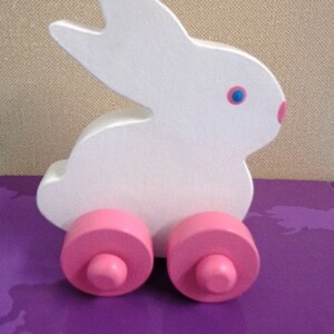 May include: A white wooden bunny toy with pink wheels. The bunny has blue eyes and a pink nose. The toy is on a purple surface. A cute and colorful toy for children.