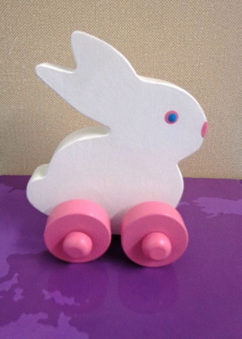 May include: A white wooden bunny toy with pink wheels. The bunny has blue eyes and a pink nose. The toy is on a purple surface. A cute and colorful toy for children.