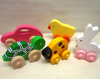 Könnte beinhalten: Ein Set aus fünf Holz-Ziehspielzeugen. Die Spielzeuge sind ein rosa Auto, eine grüne Schildkröte, ein gelber Marienkäfer, ein gelber Küken und ein weißes Kaninchen. Jedes Spielzeug hat Räder und ist in leuchtenden Farben bemalt.