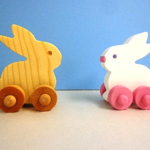May include: Two wooden toy bunnies. One is natural wood with brown wheels, the other is white with pink wheels. Both have a simple design with large ears and are on a light surface with a blue background.