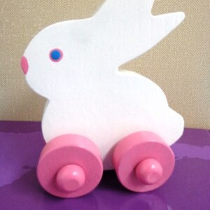 May include: A white wooden bunny toy with pink wheels and a pink nose. The bunny has blue eyes and is designed in a simple, playful style. This toy is suitable for young children.
