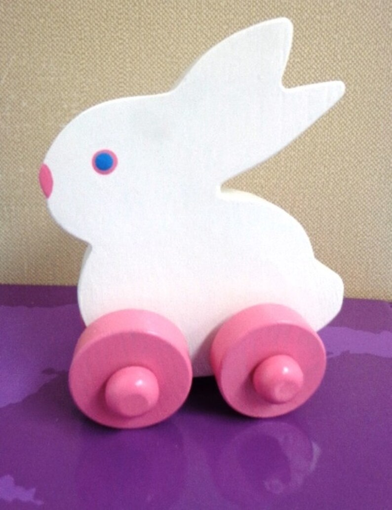 May include: A white wooden bunny toy with pink wheels and a pink nose. The bunny has blue eyes and is designed in a simple, playful style. This toy is suitable for young children.