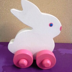May include: A white wooden bunny toy with pink wheels and a pink nose. The bunny has blue eyes and is designed in a simple, cartoonish style. This toy is suitable for young children.