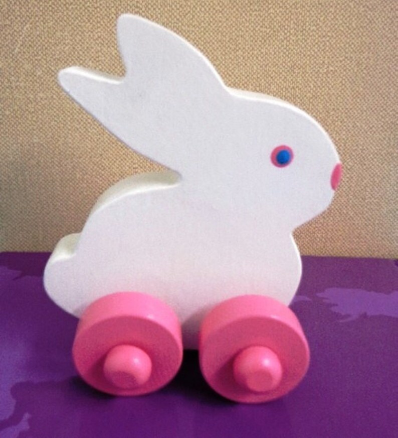 May include: A white wooden bunny toy with pink wheels and a pink nose. The bunny has blue eyes and is designed in a simple, cartoonish style. This toy is suitable for young children.