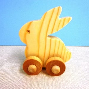 May include: A wooden toy bunny with a light yellow finish. The bunny has a simple design with two large ears and is mounted on two wooden wheels. The toy is set against a light blue background.