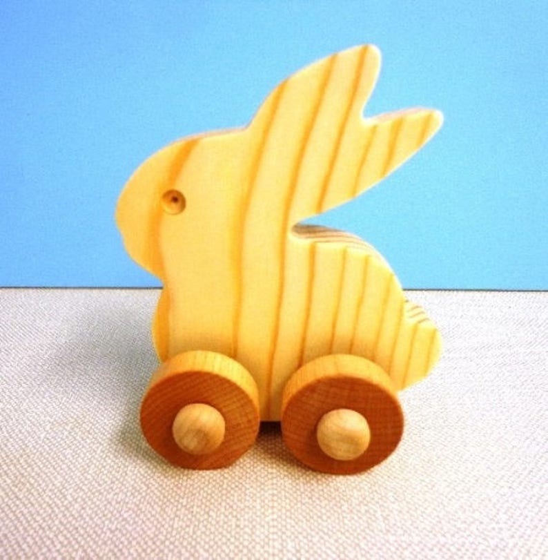 May include: A wooden toy bunny with a light yellow finish. The bunny has a simple design with two large ears and is mounted on two wooden wheels. The toy is set against a light blue background.