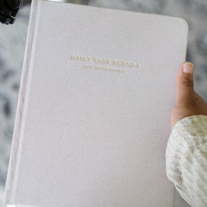 May include: A light gray daily task agenda notebook with the text "DAILY TASK AGENDA" and "JUNE PAPER MARKET" printed in gold lettering.