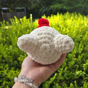 Puede incluir: Un cupcake blanco tejido a mano con una cereza roja encima. El cupcake tiene un aspecto esponjoso y texturizado y se sostiene en una mano sobre un fondo de follaje verde. El cupcake es un art&iacute;culo decorativo caprichoso.