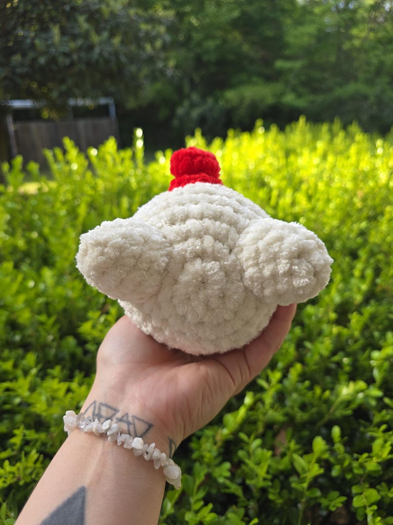 Puede incluir: Un cupcake blanco tejido a mano con una cereza roja encima. El cupcake tiene un aspecto esponjoso y texturizado y se sostiene en una mano sobre un fondo de follaje verde. El cupcake es un art&iacute;culo decorativo caprichoso.