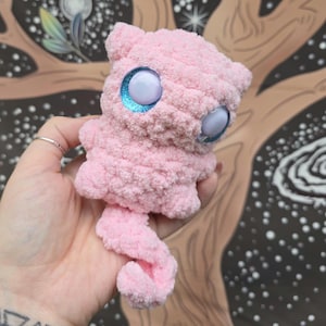 May include: A handmade pink plush creature with large, light blue eyes. The plush toy has a fluffy texture and a curled tail. The background features a tree design with a starry night sky.