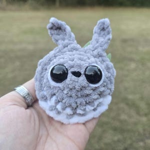 May include: A handmade gray crochet creature with large black eyes and small, upright ears. The plush toy has a white belly and a small green leaf detail. The soft, fuzzy texture is visible in this close-up shot.