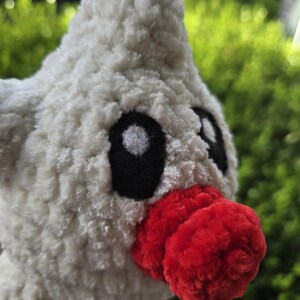Puede incluir: Un juguete de peluche hecho a mano con forma de p&aacute;jaro. El juguete es principalmente blanco con una textura esponjosa. Presenta ojos negros y un pico rojo brillante. El fondo es verde borroso, lo que sugiere un entorno al aire libre.