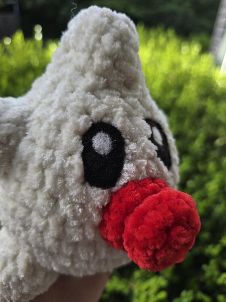 Puede incluir: Un juguete de peluche hecho a mano con forma de p&aacute;jaro. El juguete es principalmente blanco con una textura esponjosa. Presenta ojos negros y un pico rojo brillante. El fondo es verde borroso, lo que sugiere un entorno al aire libre.
