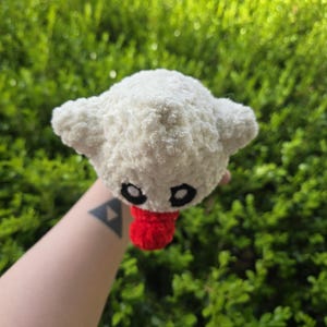 Puede incluir: Un peluche blanco hecho a mano, con ojos negros y una lengua roja. El juguete tiene dos orejas peque&ntilde;as y redondeadas y est&aacute; hecho de un material suave y texturizado. El peluche se sostiene contra un fondo de follaje verde.
