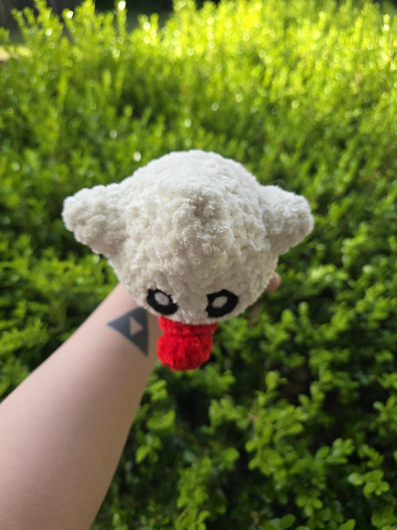 Puede incluir: Un peluche blanco hecho a mano, con ojos negros y una lengua roja. El juguete tiene dos orejas peque&ntilde;as y redondeadas y est&aacute; hecho de un material suave y texturizado. El peluche se sostiene contra un fondo de follaje verde.
