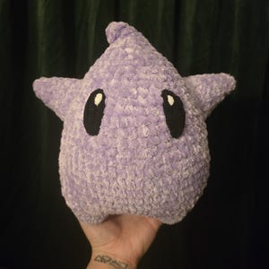 May include: A plush, lavender-colored star-shaped pillow with black oval eyes. The soft, textured pillow has two small star-shaped arms extending from the sides. The pillow is held in a hand against a dark green curtain background.