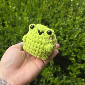 May include: A small, round, lime green crocheted frog with black eyes and a small pink blush on its cheeks. The frog has a simple, smiling expression and is made of a soft, textured yarn. The frog is a handmade item.