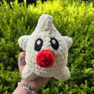 Puede incluir: Un juguete de peluche hecho a mano con forma de estrella, en hilo color crema. Presenta grandes ojos negros ovalados y una nariz redonda roja brillante. El juguete se sostiene sobre un fondo de follaje verde.