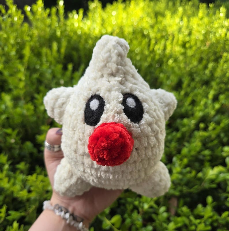 Puede incluir: Un juguete de peluche hecho a mano con forma de estrella, en hilo color crema. Presenta grandes ojos negros ovalados y una nariz redonda roja brillante. El juguete se sostiene sobre un fondo de follaje verde.
