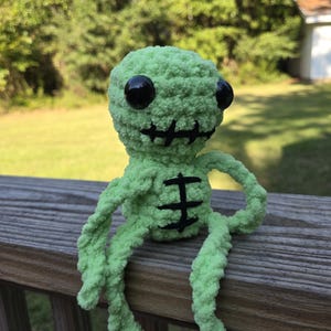 May include: A handmade, green crocheted creature with large black eyes and a stitched mouth. The plush toy has long, dangling limbs and a black stitched design on its chest. The creature is sitting on a wooden surface, with a blurred green background.
