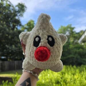 Puede incluir: Un juguete de peluche hecho a mano, en forma de estrella, tejido en blanco, con ojos negros y una nariz roja. El juguete tiene peque&ntilde;as protuberancias en forma de orejas. Se sostiene contra un fondo de follaje verde y cielo azul.