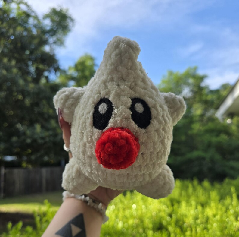 Puede incluir: Un juguete de peluche hecho a mano, en forma de estrella, tejido en blanco, con ojos negros y una nariz roja. El juguete tiene peque&ntilde;as protuberancias en forma de orejas. Se sostiene contra un fondo de follaje verde y cielo azul.