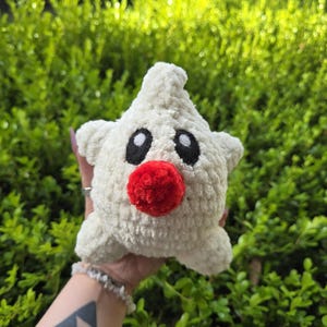 Puede incluir: Un juguete de peluche hecho a mano con forma de estrella, en hilo de color crema. Presenta ojos ovalados negros y una nariz redonda de color rojo brillante. El juguete es suave y tierno, perfecto para ni&ntilde;os o como objeto decorativo.