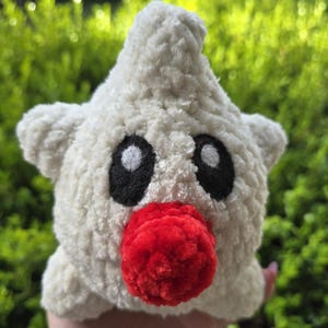 Puede incluir: Un juguete de peluche hecho a mano con forma de estrella, de textura blanca y esponjosa. Presenta grandes ojos negros, una nariz roja y peque&ntilde;as protuberancias en forma de orejas. El fondo es borroso y verde.