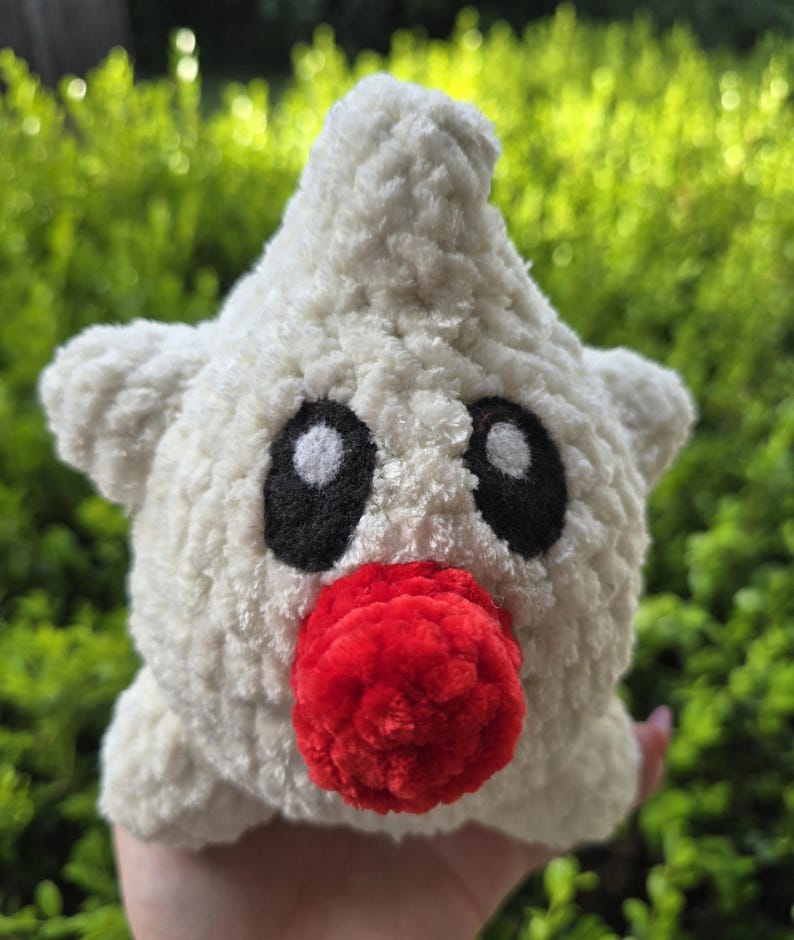 Puede incluir: Un juguete de peluche hecho a mano con forma de estrella, de textura blanca y esponjosa. Presenta grandes ojos negros, una nariz roja y peque&ntilde;as protuberancias en forma de orejas. El fondo es borroso y verde.