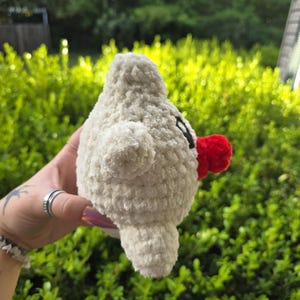 Puede incluir: Una criatura blanca de peluche hecha a mano, con forma de p&aacute;jaro, con un pico rojo y peque&ntilde;os ojos negros. El juguete suave y texturizado tiene peque&ntilde;as alas y un cuerpo redondeado. El fondo es una vista borrosa de follaje verde.