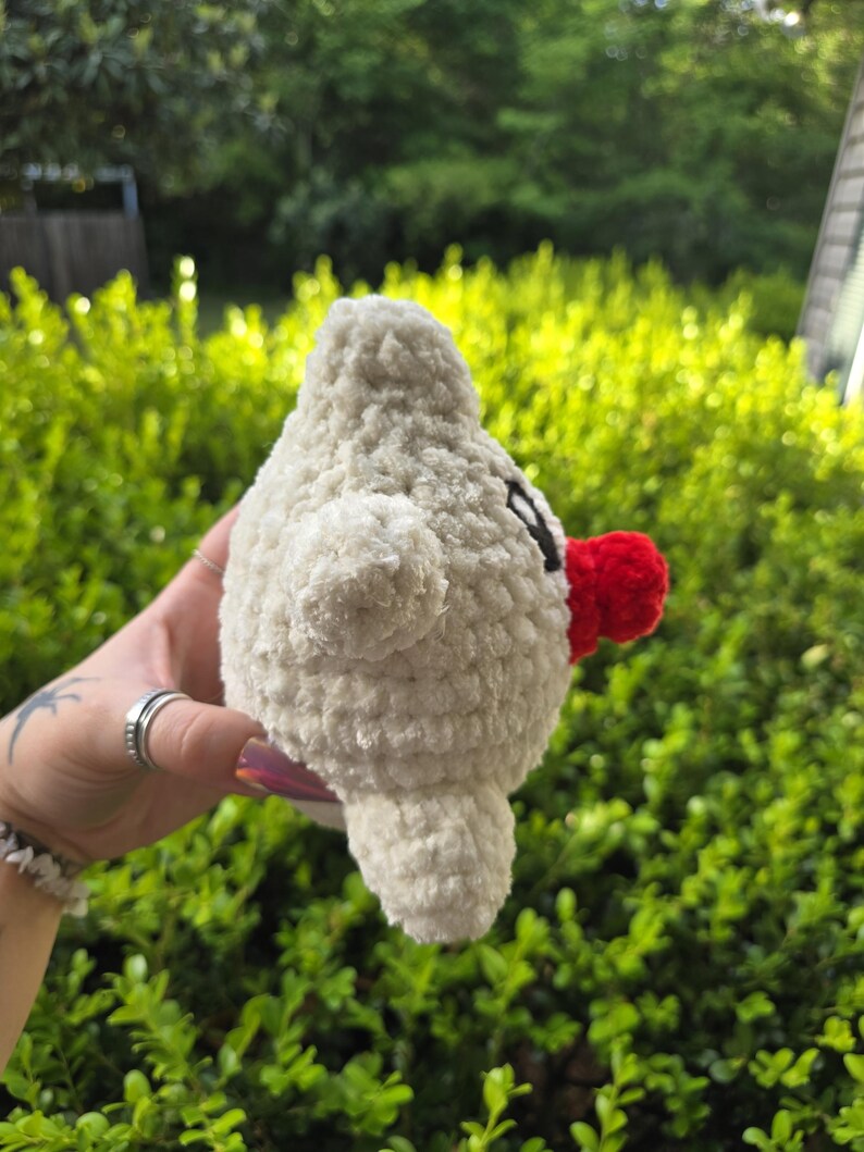 Puede incluir: Una criatura blanca de peluche hecha a mano, con forma de p&aacute;jaro, con un pico rojo y peque&ntilde;os ojos negros. El juguete suave y texturizado tiene peque&ntilde;as alas y un cuerpo redondeado. El fondo es una vista borrosa de follaje verde.