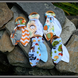 May include: Five wooden teethers with fabric loops. Each teether has a different animal print and a soft, white fabric on the back. The teethers are arranged on a bed of gray rocks.