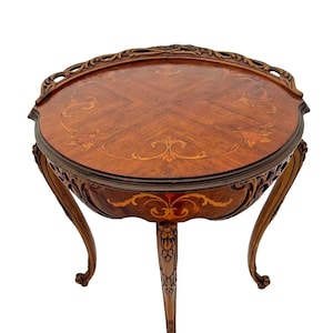May include: A round, wooden table with intricate marquetry inlay on the top. The table has three legs with carved details and a scalloped edge.