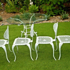 Pode incluir: Conjunto de p&aacute;tio de ferro fundido branco com uma mesa redonda de vidro e quatro cadeiras. As cadeiras t&ecirc;m um design de treli&ccedil;a e um encosto curvo.