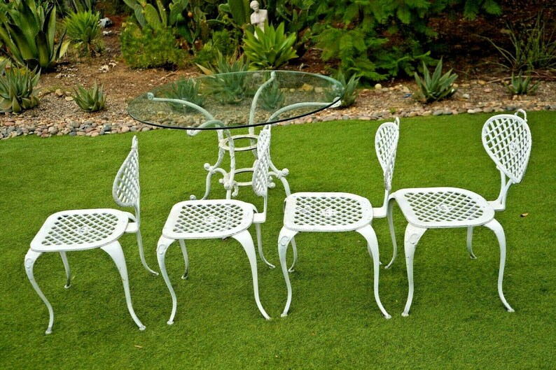 Pode incluir: Conjunto de p&aacute;tio de ferro fundido branco com uma mesa redonda de vidro e quatro cadeiras. As cadeiras t&ecirc;m um design de treli&ccedil;a e um encosto curvo.