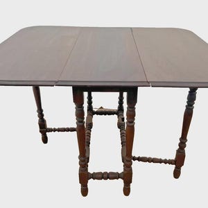 May include: A dark brown, rectangular drop-leaf table with rounded corners. The table has a three-section top and ornate, turned legs. The table is approximately 36 inches wide and 48 inches long when fully extended.