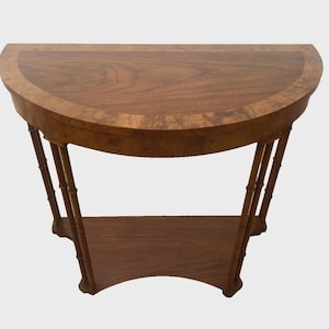 May include: A half-moon shaped wooden table with a dark brown finish. The table has a lower shelf and is supported by bamboo-style legs. The tabletop has a lighter wood border. A classic piece of furniture.
