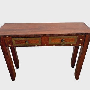 May include: A small, wooden console table with two drawers. The table is a rich brown color with brass accents around the drawers and decorative studs. The legs are angled, and the table has a simple, elegant design.
