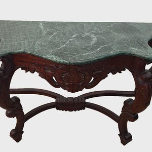 May include: A semi-circular console table featuring a dark brown carved wooden base with ornate scrollwork and a smooth, polished green marble top with white veining. The structure includes four curved legs connected by a central stretcher.