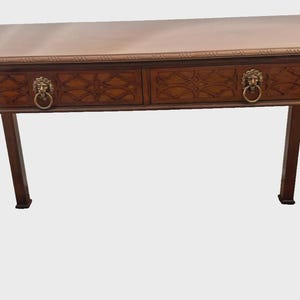 May include: A rectangular wooden desk with a smooth top surface and two drawers. Each drawer features a carved lattice pattern and a brass lion head pull handle. The desk stands on four straight legs with decorative carved brackets at the top.